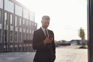 Kendine güvenen olgun iş adamı ofis binasının yanında dururken akıllı telefon kullanıyor.