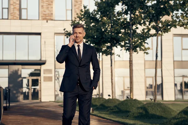 Confiado hombre de negocios maduro hablando por teléfono móvil mientras camina cerca del edificio de oficinas al aire libre —  Fotos de Stock