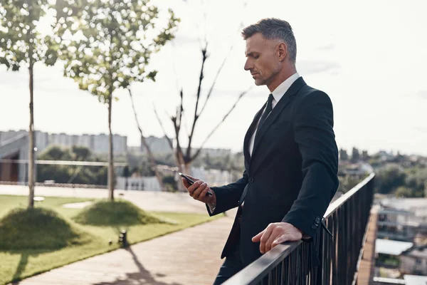 Důvěryhodný zralý podnikatel se dívá na svůj chytrý telefon, zatímco stojí venku — Stock fotografie