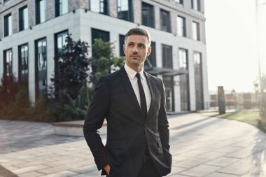 Takım elbiseli, kendine güvenen olgun bir adam ofis binasının yanında duruyor ve ellerini cebinde tutuyor.