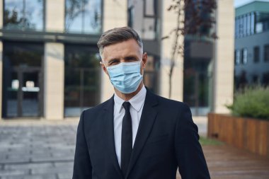Koruyucu yüz maskesi takmış, dışarıda kameraya bakan kendinden emin bir iş adamı.