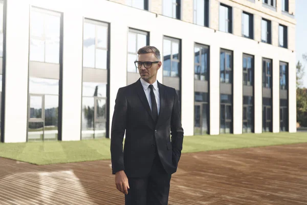 Fiducioso uomo d'affari maturo in occhiali da vista guardando lontano mentre in piedi di fronte a edificio per uffici — Foto Stock