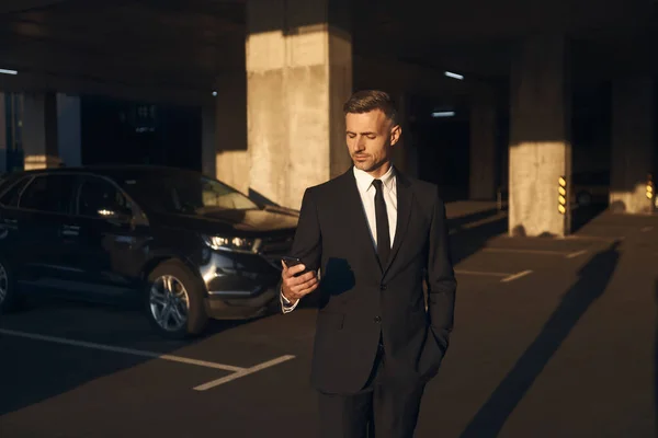 Confident mature businessman looking at his mobile phone while standing on parking lot — Stock Photo, Image
