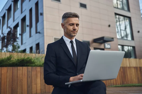 Selbstbewusster, reifer Geschäftsmann arbeitet am Laptop, während er im Freien in der Nähe eines Bürogebäudes sitzt — Stockfoto