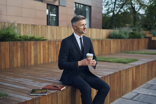 Uvolněný zralý podnikatel pije kávu, zatímco sedí venku v blízkosti kancelářské budovy — Stock fotografie