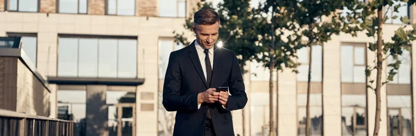 Confident mature businessman looking at mobile phone while walking near office building outdoors — Stock Photo, Image