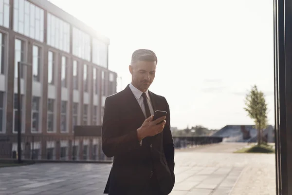 Důvěryhodný zralé podnikatel pomocí chytrého telefonu, zatímco stojí v blízkosti kancelářské budovy venku — Stock fotografie