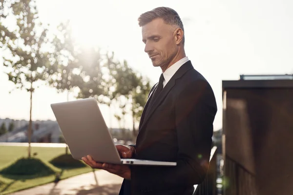 Důvěryhodný zralý podnikatel pracující na notebooku, zatímco stojí venku — Stock fotografie