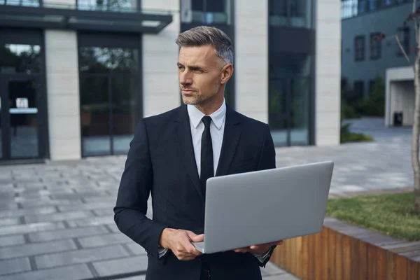 Důvěryhodný zralý podnikatel pracující na notebooku, zatímco stojí venku v blízkosti kancelářské budovy — Stock fotografie