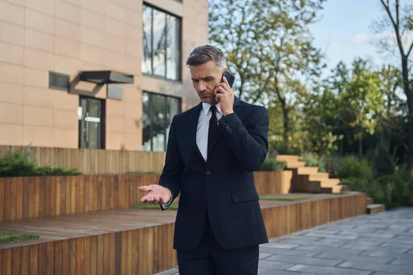 Empresário maduro confiante falando no telefone celular enquanto está ao ar livre perto do prédio de escritórios — Fotografia de Stock