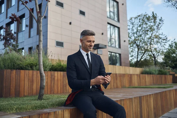 Αυτοπεποίθηση ώριμος επιχειρηματίας χρησιμοποιώντας έξυπνο τηλέφωνο, ενώ κάθεται σε εξωτερικούς χώρους κοντά στο κτίριο γραφείων — Φωτογραφία Αρχείου