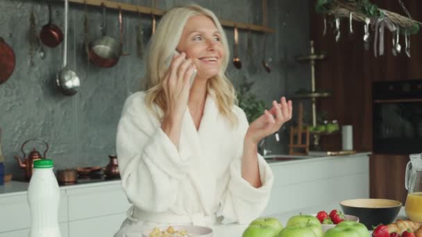 Mujer madura hablando por teléfono móvil mientras desayuna en la cocina — Vídeo de stock