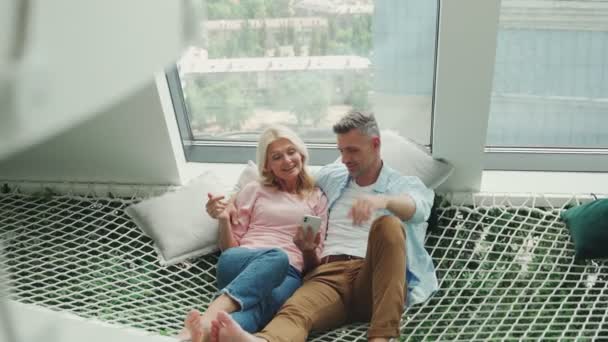 一对成熟的夫妇一起在家里的吊床上一边检查智能手机一边放松 — 图库视频影像