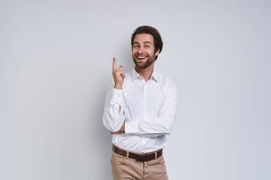 Beyaz gömlekli yakışıklı genç adam kameraya bakıyor ve gri arka planda dururken fotokopi alanını işaret ediyor.