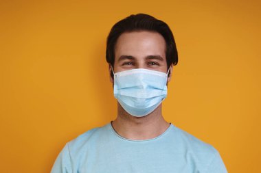 Sarı arka planda dikilirken kameraya bakan koruyucu maskeli bir gencin portresi