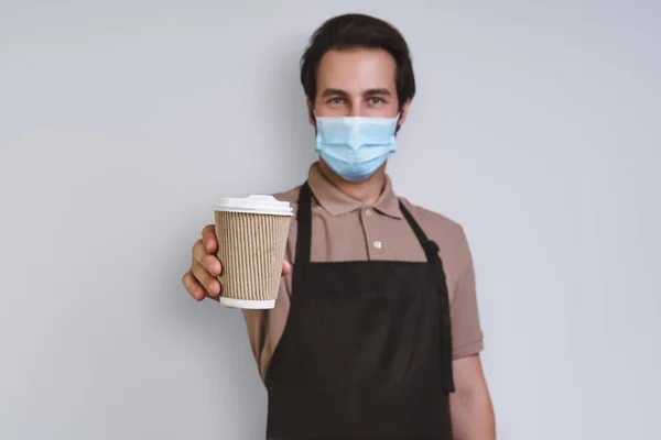 Confident young man in apron wearing protective face mask and holding coffee cup while standing against gray background — Stock Photo, Image