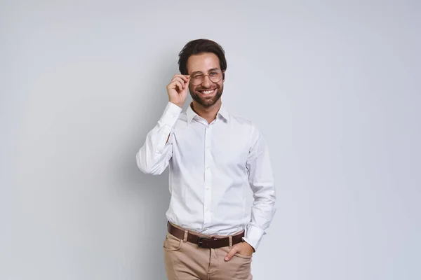 Knappe jongeman in wit shirt die naar de camera kijkt en zijn bril aanpast terwijl hij tegen een grijze achtergrond staat — Stockfoto