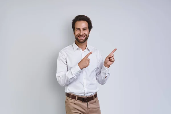 Schöner junger lächelnder Mann in weißem Hemd, der vor grauem Hintergrund in die Kamera blickt und wegzeigt — Stockfoto