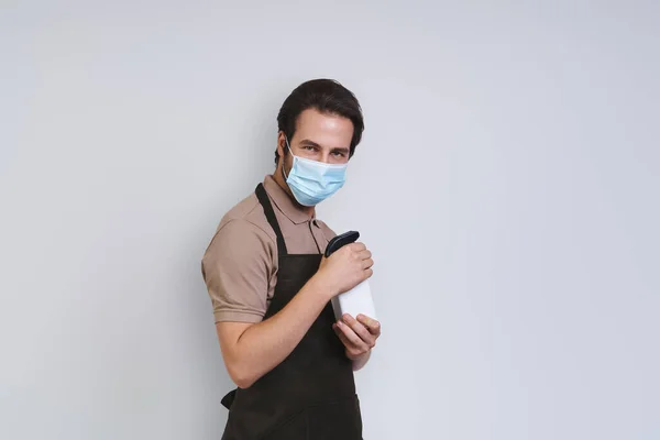 Handsome young man in apron wearing protective face shield and holding cleaning spray while standing against gray background — Stock Photo, Image