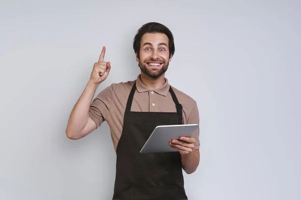Glada ung man i förklädet visar sin digitala tablett och gester när du står mot grå bakgrund — Stockfoto