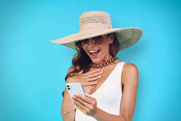 Surprised young woman in elegant hat looking at her mobile phone while standing against blue background — Stock Photo, Image