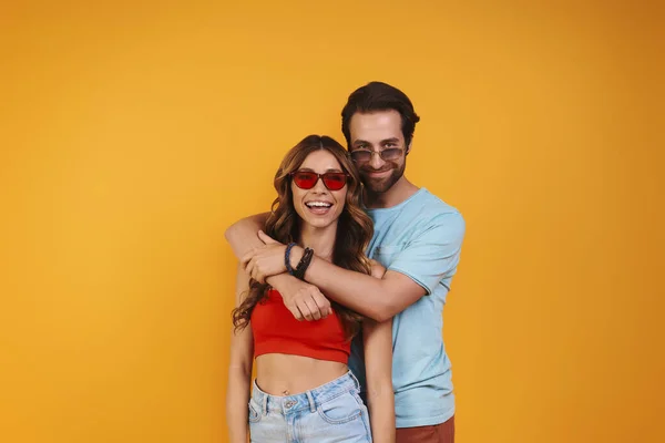 Hermosa pareja joven en gafas de vista abrazándose mientras está de pie sobre el fondo amarillo —  Fotos de Stock