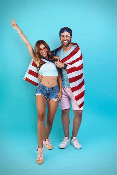 Pełna długość pięknej młodej pary pokrytej amerykańską flagą uśmiechając się stojąc na niebieskim tle — Zdjęcie stockowe