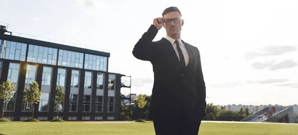 Confiado hombre de negocios maduro ajustando sus gafas mientras está parado al aire libre — Foto de Stock