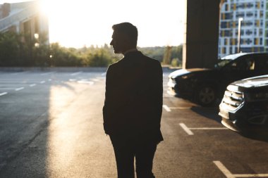 Formalite icabı giyinmiş bir adam otoparkta duruyor ve arka planda gün batımı var.