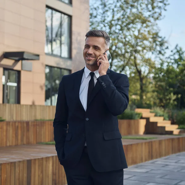 Confiado hombre de negocios maduro hablando por teléfono móvil mientras está parado al aire libre cerca del edificio de oficinas — Foto de Stock