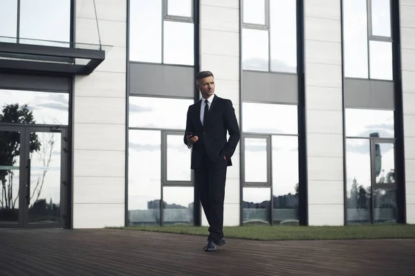 Fiducioso uomo d'affari maturo che tiene il telefono cellulare mentre cammina davanti all'edificio dell'ufficio — Foto Stock