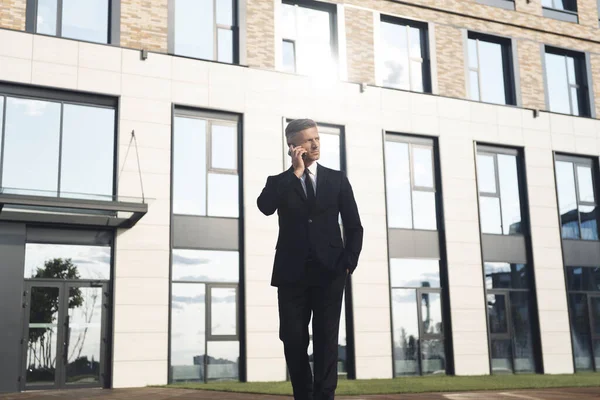 Empresário maduro confiante falando no telefone celular enquanto caminha na frente do prédio de escritórios — Fotografia de Stock