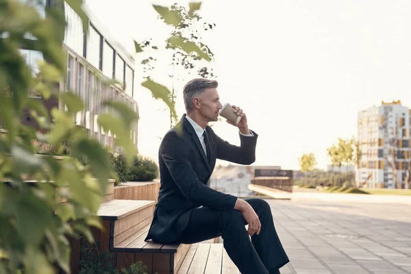 Ontspannen volwassen zakenman drinken van koffie tijdens het zitten buiten in de buurt van kantoorgebouw — Stockfoto