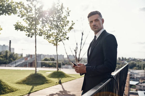 Fiducioso uomo d'affari maturo che trasporta smartphone e distoglie lo sguardo mentre si trova all'aperto — Foto Stock