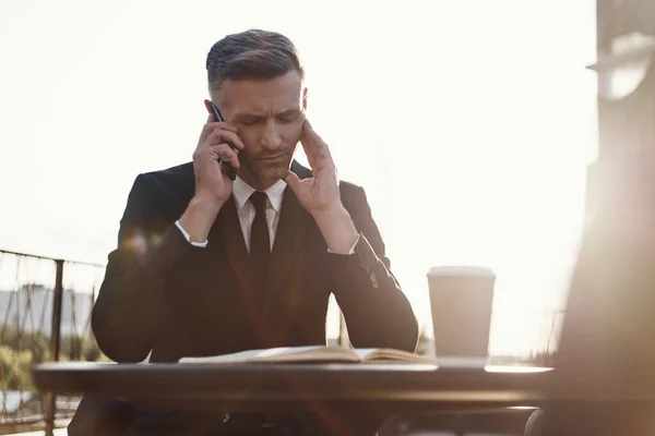 Concentrated mature businessman talking on mobile phone and making notes while sitting in cafe outdoors — Stock Photo, Image