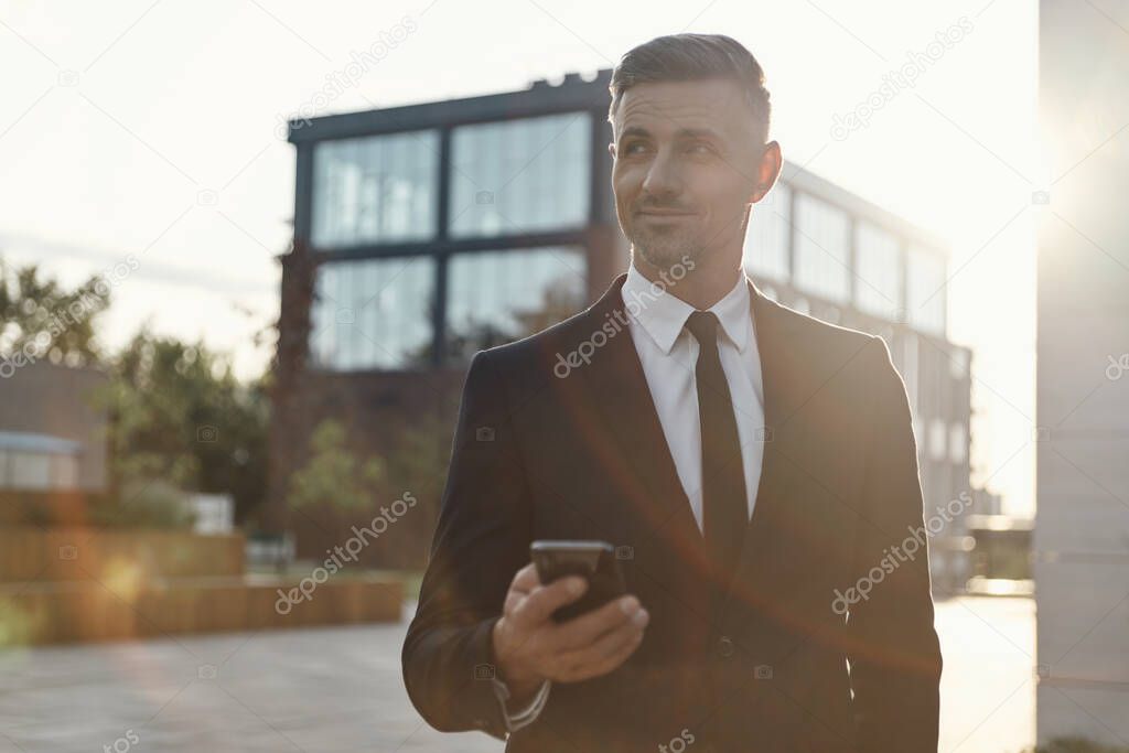 Confident mature businessman holding mobile phone while standing near office building outdoors