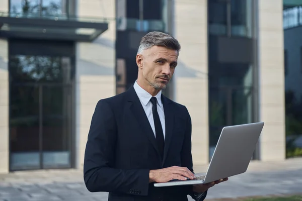 Homme d'affaires mature confiant travaillant sur ordinateur portable tout en se tenant à l'extérieur près de l'immeuble de bureaux — Photo