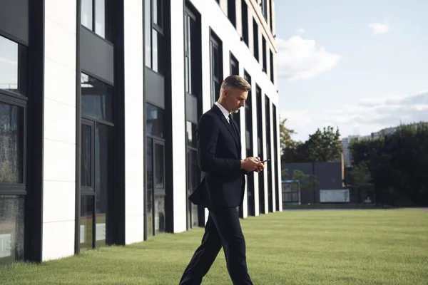Empresário maduro confiante segurando telefone celular enquanto caminhava perto do prédio de escritórios — Fotografia de Stock