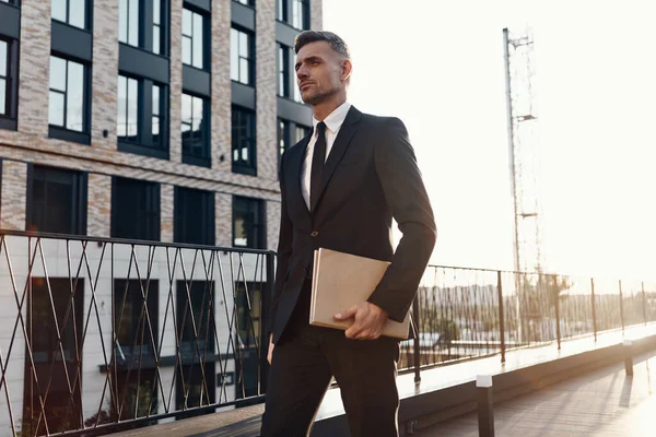 Pewny siebie dojrzały mężczyzna w pełnym garniturze niosący laptopa podczas spaceru przed budynkiem biurowym — Zdjęcie stockowe