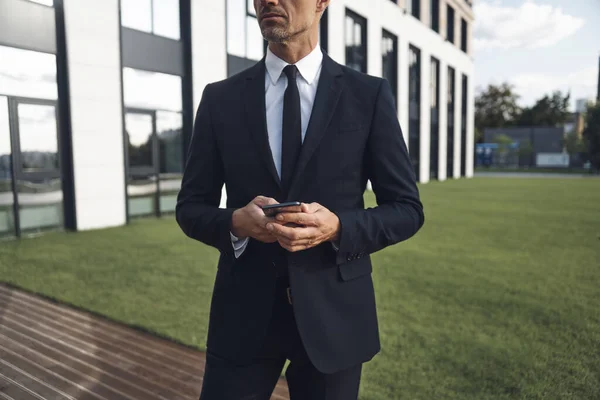 Confident mature businessman holding mobile phone while standing near office building — Stock Photo, Image