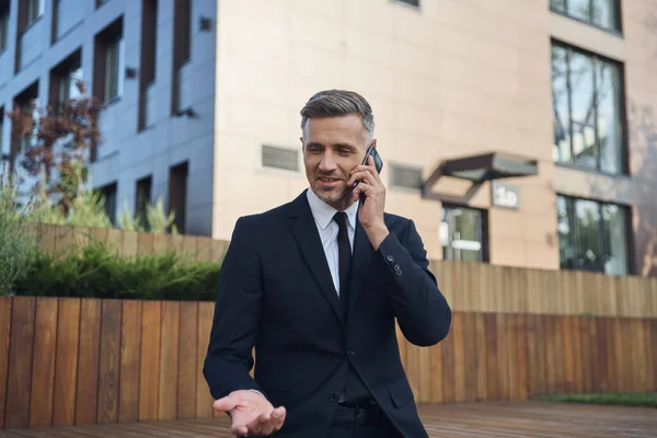 Empresário maduro confiante falando no telefone celular enquanto sentado ao ar livre perto do prédio de escritórios — Fotografia de Stock