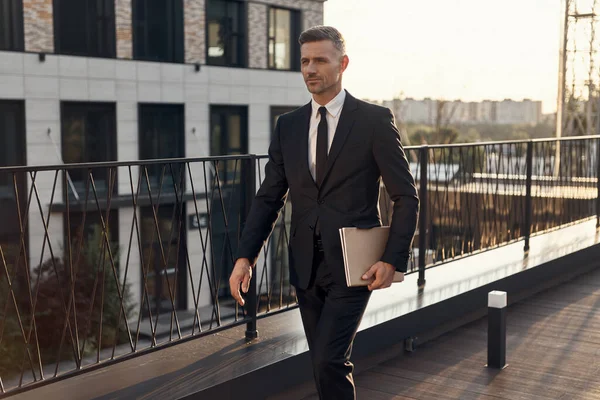 Homem maduro confiante em terno completo carregando laptop enquanto caminhava pelo terraço do centro de negócios ao ar livre — Fotografia de Stock