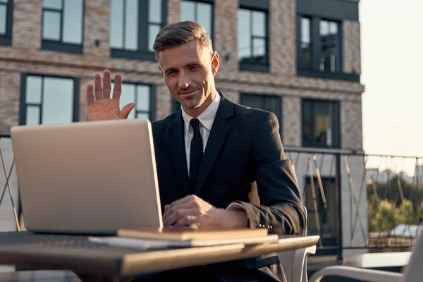 Joyeux homme d'affaires mature ayant appel vidéo sur ordinateur portable tout en étant assis dans un café à l'extérieur — Photo