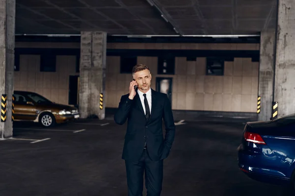 Confident mature businessman talking on mobile phone while walking by parking lot — Stock Photo, Image