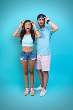 Mavi arka planda dikilirken kameraya bakan tam boy şaşırmış genç çift.