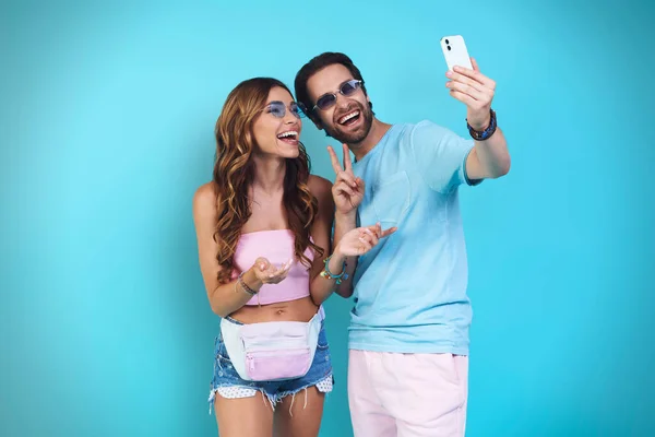 Bonito jovem casal sorrindo fazendo selfie enquanto está de pé contra o fundo azul — Fotografia de Stock