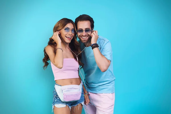 Hermosa joven sonriente pareja en auriculares escuchando música mientras está de pie sobre fondo azul —  Fotos de Stock