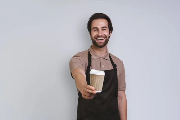 Cheerful young man in apron holding coffee cup and smiling while standing against gray background — Stock Photo, Image