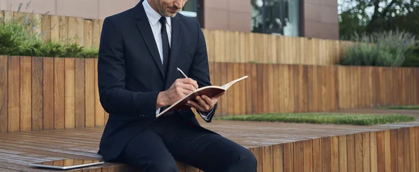 Pewny siebie dojrzały biznesmen robi notatki siedząc na zewnątrz w pobliżu biurowca — Zdjęcie stockowe