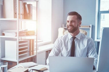 Gömlekli ve kravatlı mutlu genç adam ofiste otururken bilgisayarla çalışıyor ve gülümsüyor.